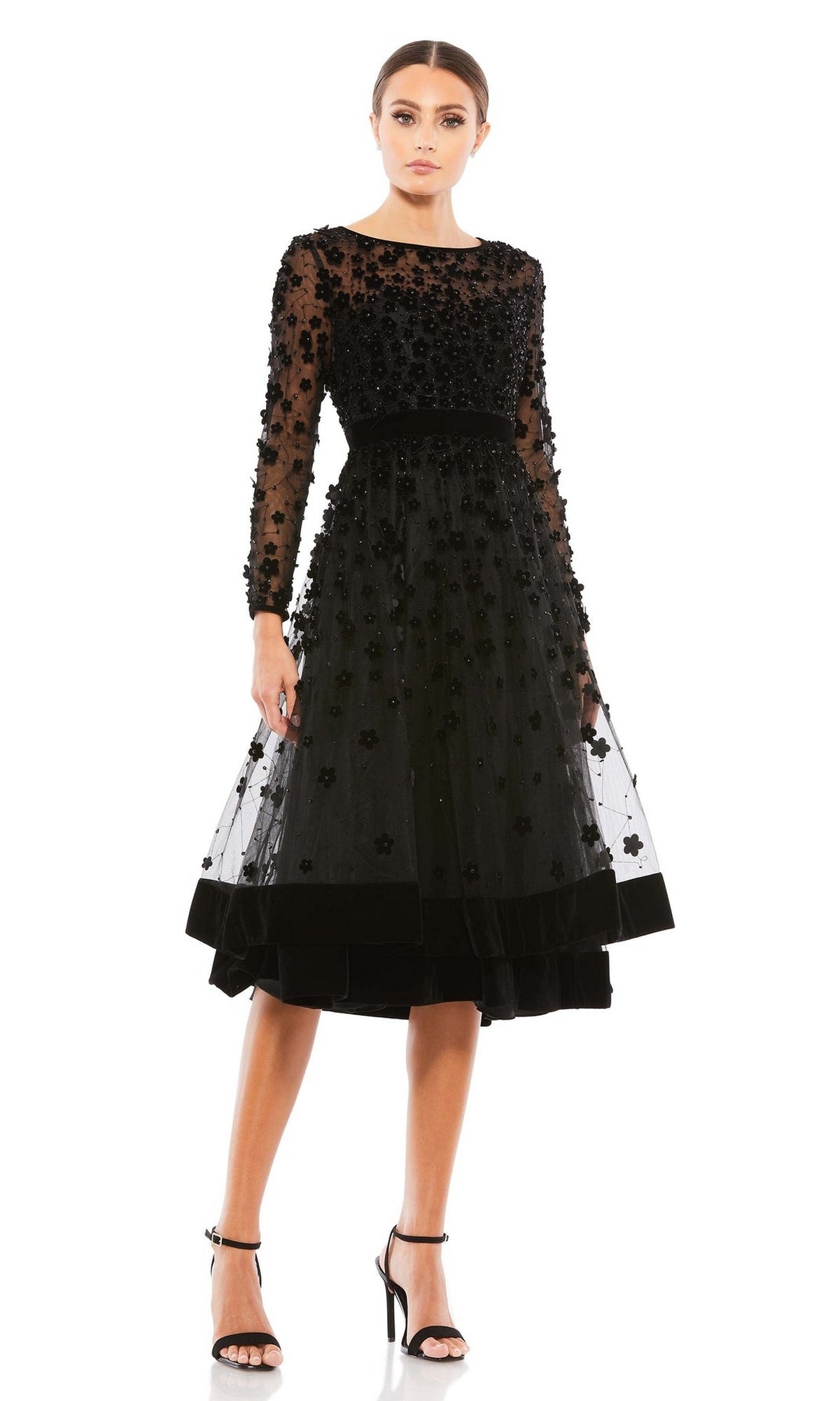 A woman stands against a white background wearing the Mac Duggal 67007 knee-length wedding-guest dress in black with floral embellishments, sheer long sleeves, and paired with black heels.